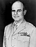 Head and shoulders of a balding middle-aged white man wearing a light-colored military jacket with three stars on the shoulder and four rows of ribbon bars and a winged pin on the left breast.