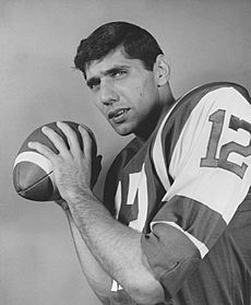 Black and white publicity still of Namath, in pads and jersey with no helmet, holding football over his shoulder as if to throw it (1965).
