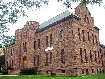 A large brown stone building with castle-like towers and a pointed roof with step-like features on the end. Hanging from its right side is a banner that says "Lake Plains YMCA"