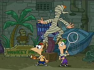Two cartoon boys stand beside each other looking in opposite directions. A cartoon girl with red hair stands clumsily on their heads, wrapped in raggy toilet paper. Behind them are pipes, a gray brick wall, and signs.