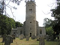 Front view of St Peter's church showing the late Anglo-Saxon flint round tower; Anglo-Saxon double bell opening; 14th century parapet, stringcourse and gargoyles. Set within a large churchyard with trees to both sides.