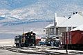 Winter photograph of the Nevada Northern Railway, East Ely Yards. A steam engine is heading a short train in front of the passenger depot, with mountainsides rising out of the picture in the far background.