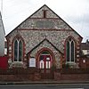 Front view of a tiny, plain chapel of irregular flintwork and red brick dressings. A dark stone plaque below a slit window near the roofline reads "WESLEYAN CHAPEL".  An entrance porch is flanked by two wide, low-set windows with pointed arches.