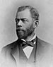Head and shoulders of a white man with a full beard, wearing a dark suit coat, vest, and bow tie.
