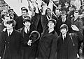 Black and white picture of the Beatles waving in front of a crowd with an set of aeroplane steps in the background 