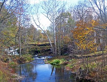 A creek with some waterfalls over an old dam in the distance on a clear autumn day. There is a house on the right obscured by the trees
