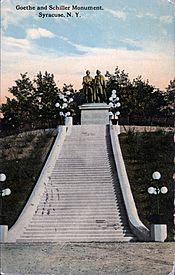 Photograph of an old color postcard. There is printing at the top left of the postcard that says "Goethe and Schiller Monument, Syracuse, N.Y." The picture shows a statue of two men on a stone pedestal. The statue is at the top of a slope, and is approached by a long, wide stairway. There are several lighting fixtures on each side of the stairway, and there are formal plantings of shrubbery on both sides. There are trees behind the statue.