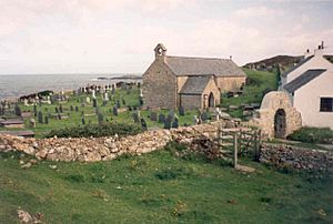 A small windowless church in a churchyard, with the sea visible in the distance. A stone arch is visible over the path leading into the churchyard.