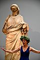 An actress performing a play. She wears a laurel wreath and stands in front of a statue of a woman from the Mausoleum at Halicarnassus. Room 21, The British Museum, London