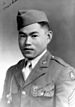 Head and shoulders of a young man wearing a garrison cap and a military jacket with three chevrons on the upper left sleeve and a whistle hanging from a chain attached to his right shoulder. In the top left corner of the photo is written "To Mom & Dad" and in the lower right "your son! Kazuo".
