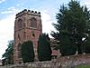 A red sandstone crenellated church tower in front of which are yew trees and a wall