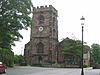 A sandstone church seen from the west with the tower prominent. To the left is a tree and part of a red car, and to the right is a lamppost