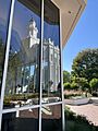 A glass window from the visitors center reflects the St. George Temple, with tall white parapets and a domed octagonal spire. Various foliage is seen off to the side.