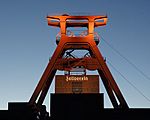 A orange metal tower with several flywheels above a building with Zollverein written in golden gothic script letters.