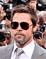 A Caucasian male, who is wearing aviator sunglasses, has light brown hair and a short brown beard. He wears a grey suit jacket, white shirt, and grey tie. Behind him are people with single-lens reflex cameras.