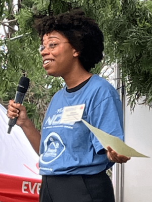 A smiling young Black woman outdoors, holding a microphone and a paper, wearing glasses and a blue t-shirt with a name badge