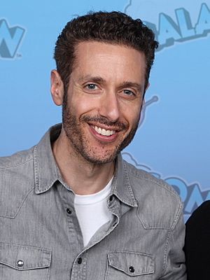 Paulo Costanzo with stubble wearing a faded grey denim shirt with white t-shirt underneath, standing in front of blue background, grinning at camera
