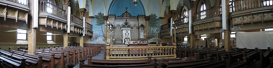 A wide, panoramic view of a synagogue sanctuary can be seen. Three rows of wooden pews lead to the front of the room; the middle row is interrupted by a raised square wooden platform, surrounded by a heavy wooden railing with lights on each corner. At the front of the room is a large wooden ark, surrounded on three sides by painted scenes of buildings and trees. At the sides of the room are balconies with heavy wooden railings, interrupted by large columns.