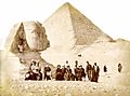 A large group of men and women are gathered below the head of the Sphinx with the Great Pyramid looming behind