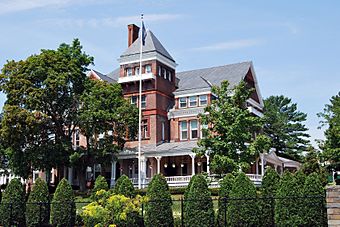 An ornate brick house with a pyramid-roofed tower on the front seen from slightly below. In front is a flagpole and some tall trees, with shrubbery and a chain link fence at the bottom of the image, closer to the camera.