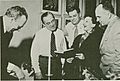 Four middle-aged men are gathered around to listen to a woman sing from a book