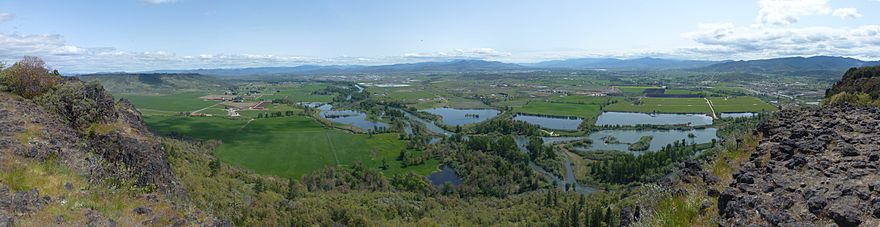 A river, seen from above, winds through flat farmland and a series of ponds on both sides of the stream. Buildings are scattered here and there across the landscape under a cloudy sky.
