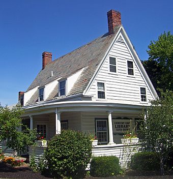 View of side of house, with flowers and shrubbery in front and a sign saying "Schenectady County Public Library,  Scotia Branch" on wall behind porch.
