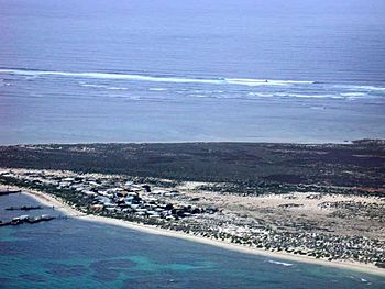 View across a sandy strip of land, with ocean on both sides. The land nearest the observer is mostly sandy, and has many buildings on it, and some jetties. The land furthest from the observer is thickly vegetated. The ocean in the foreground is blue-green, with dark patches indicating areas of coral reef. The ocean in the background is light blue. There is a line of breakers in the distance.