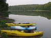 One blue and two yellow kayaks on the shore of a lake, which reflects surrounding pine trees