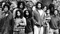 A black and white photo of group of around a dozen people standing outside. Everyone in the photo is black and has the same style of dreadlocks. The ages of the group vary, from a baby being held in someone's arms to a man who could be in his twenties or thirties.