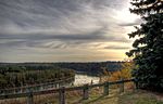 A wide river winds by a fenced overlook opposite a forest. The setting sun illuminates a partly cloudy sky.