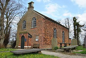 A small brick church seen from the southwest with a small porch, a bellcote, and large round-headed windows