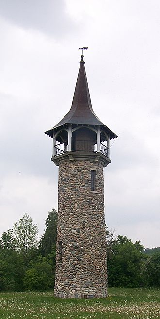 In the background is a grey, overcast sky above a canopy of trees. In the foreground is a grass field with numerous dandelions display seed heads, in the middle of which rises a tower of earth-tones multi-coloured stones. At the top of the tower is an observation deck ringed by an iron railing, each section of which is supported by end columns painted white that also support the roof structure. The copper roof is a concave structure peaking at a point, topped with an ornamental weather vane shaped like an 1800s Conestoga wagon.