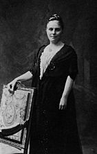 A posed formal black-and-white photograph of a woman standing behind a chair upon which she is resting her hand