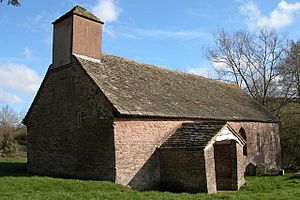A small, simple, stone church with a stone slate roof, a bellcote with a pyramidal roof, a porch and small windows, seen from the southwest