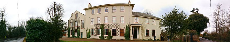 Panorama of a three-story stucco rendered building in a plain Georgian style, with bushes and trees on each side where a boundary wall shelters the gravelled garden from a narrow lane.