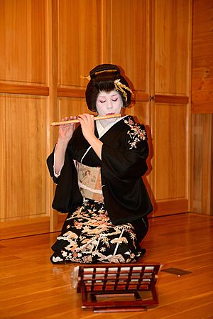 A woman wearing a black kimono, white makeup and a traditionally-styled wig sat kneeling and playing the flute.