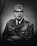 Head and torso of a man sitting with his arms folded on his lap, wearing a garrison cap, horn-rimmed glasses, and a military jacket. The jacket's left breast is completely covered in ribbon bars and medals.