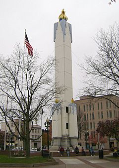 A tall white structure resembling a candle with a yellow fiberglass flame on the top sits in the center of a town square-like area. At the base of the structure are two smaller, identical-looking candle-life structures. A line of people sit on a small brick wall running in front of the structure. In the background are small buildings and a flagpole with an American flag. In the foreground are two trees without leaves.