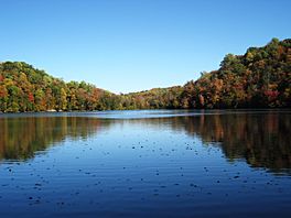 Photograph of a still lake; behind the lake are forested cliffs whose trees have autumn colors.