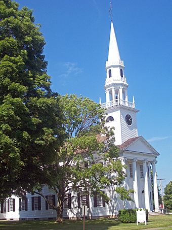 A white building with a pointed clock tower and colonnaded front on the right partially obscured by trees on the left