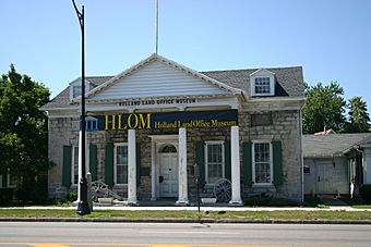 A light brown stone building with a pointed roof, green shutters and white columns on the front. In front of it are a sidewalk, a blue marker with gold writing, and a portion of street. Black letters above the front columns read "Holland Land Office Museum", which is also on a banner across the front.