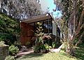 Photograph of the Eames House, a boxy building with a face of rectangles of various colors and clear glass, shaded by tall eucalyptus trees.