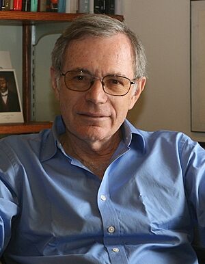 A grey-haired bespectacled man wearing a light blue shirt and sitting on a chair behind a desk; behind him is a bookshelf and a wall mounted with certificates and awards