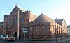 A wide terracotta-coloured brick building in two parts: to the left, a two-storey structure with three lancet windows above the arched entrance porch, spirelets and recessed parts with paired and single lancets, all topped with a triangular roof with vertical recessed sections; and to its right, a rounded single-storey section with single lancets separated by buttresses.