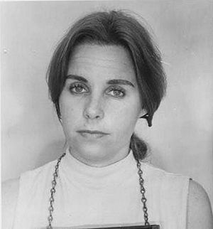 A young white woman with straight hair parted center and dressed back to the nape; she is wearing a light-colored sleeveless top with a mock turtleneck, and the chain of a police number board is visible around her neck, because this is a mug shot.