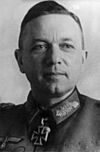 The head of a man, shown in semi-profile. He wears a military uniform with a military decoration in shape of an iron cross displayed at the front of his shirt collar. His hair is dark and combed to back.