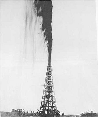 Black-and-white photograph of an oil gusher derrick with a gusher of oil shooting from the top