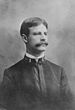 Head and shoulders of a white man with a wide mustache and carefully combed hair wearing a plain military jacket with a single bar on each side of the collar.