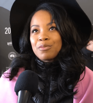 Image of actress Chanté Adams. She has brown skin, long curly hair, and is wearing a black hat.